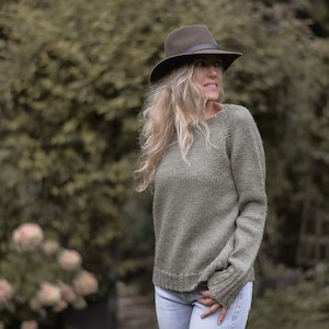 May include: A woman wearing a green knitted jumper with a brown fedora hat. The jumper has a relaxed fit and long sleeves. The woman is standing in front of a green background.