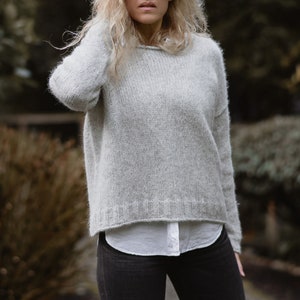 May include: A light grey knitted jumper with a relaxed fit and long sleeves. The jumper is made of a soft, fuzzy yarn and has a slightly cropped hem.