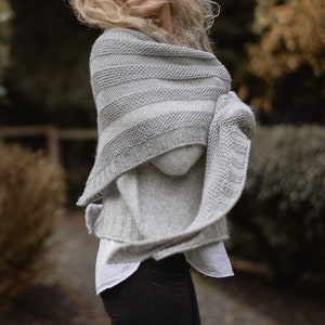 May include: A gray knitted shawl with a striped pattern. The shawl is draped over a person's shoulders and is worn with a white shirt and black pants.