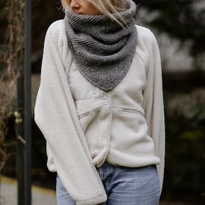 May include: A woman wearing a white fleece jacket with a gray knitted scarf. The scarf is tied around her neck and hangs down her chest.