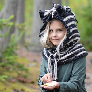 May include: A young girl wearing a black and white striped knit hooded scarf with a furry top. She is kneeling in a forest setting and holding a leaf in her hand.