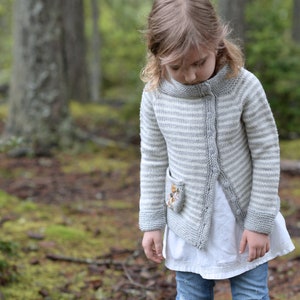 May include: A young person wearing a gray and white striped cardigan with a small embroidered pocket. The cardigan has a button closure and is worn over a white shirt and blue jeans.