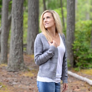 Op de afbeelding: Een vrouw draagt een grijze cardigan met een wit tanktop en blauwe jeans. Ze staat in een bos met bomen op de achtergrond.