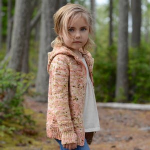 May include: A young girl wearing a pink and white hooded cardigan with a speckled pattern. The cardigan has a long sleeve and a relaxed fit. The girl is standing in a woodland setting.