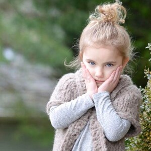 May include: A young girl wearing a brown knitted vest with pockets and a grey long-sleeved shirt. The vest has a cowl neck and is made with a chunky yarn. The girl is standing in front of a green bush.
