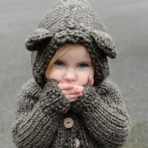 Peut inclure: Un cardigan en tricot marron avec une capuche et une fermeture à boutons. La capuche a deux petites oreilles sur le dessus. Le cardigan est porté par un enfant aux cheveux blonds.
