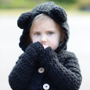 Op de afbeelding: Een zwarte gehaakte hooded cardigan met berenoren, met houten knopen en een gerimpelde donkerblauwe jurk eronder. De tekst "The Velvet Acorn" is zichtbaar onderaan de afbeelding.