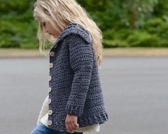 May include: A gray crocheted cardigan with a hood and button closure. The cardigan is worn by a child and paired with blue jeans and brown boots. The text "The Velvet Acorn Designs" is visible in the image.