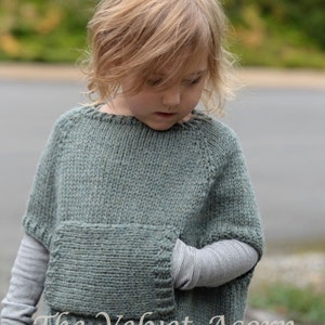 May include: A child wearing a green knitted poncho with a large pocket on the front. The poncho has a loose fit and is made with a soft, textured yarn. The child is wearing blue jeans and has blonde hair.