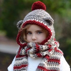 Może przedstawiać: Zestaw czapki i szalika z dzianiny w kolorze szarym i czerwonym w paski, z pomponem na czapce. Czapka ma uszy niedźwiedzia.