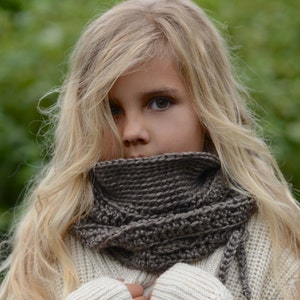 May include: A young person with long blonde hair wearing a cream-colored sweater and a dark brown knitted cowl. The cowl is wrapped around the neck and has a textured appearance. The sweater has a ribbed knit design. The Velvet Acorn is printed on the bottom of the image.