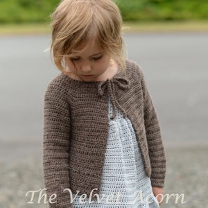 May include: A brown crocheted cardigan with a tie at the neckline. The cardigan is worn over a white crocheted dress. The text "The Velvet Acorn Designs" is visible at the bottom of the image.