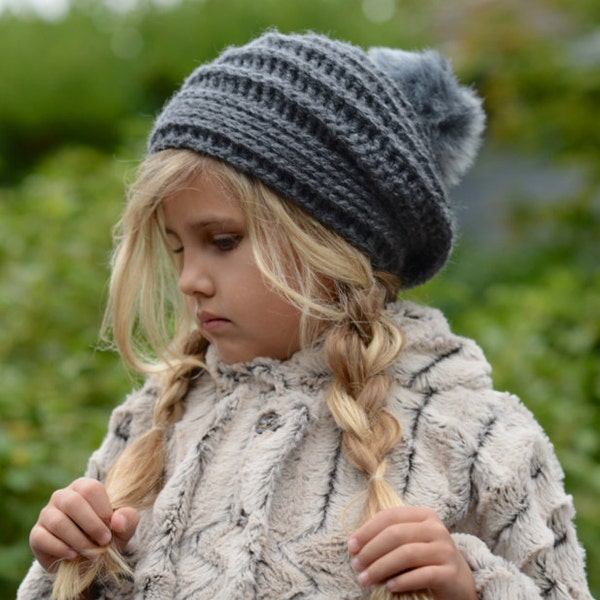 Peut inclure: Une jeune fille portant un bonnet en crochet gris avec un pompon en fausse fourrure et un poncho en fausse fourrure beige. Le poncho a un motif en chevron et la fille tient ses tresses.
