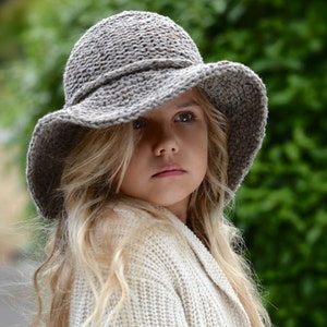 以下が含まれることがあります： グレーのクロシェのつば広帽子とクリーム色のニットカーディガンを着た若い人。帽子はテクスチャのある表面とわずかにたるんだクラウンを持っています。