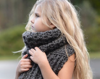 Puede incluir: Una niña pequeña con una bufanda de cuello de tortuga de ganchillo gris. La bufanda está hecha con un patrón de puntadas texturizadas. La niña tiene el pelo largo y rubio y mira hacia un lado. El texto "The Velvet Acorn Designs" es visible en la parte inferior de la imagen.