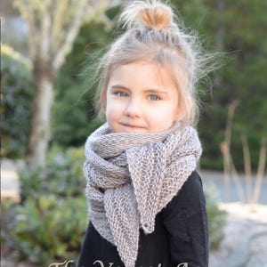 May include: A young girl wearing a grey knitted scarf with a black long-sleeved shirt. The scarf is tied around her neck and the ends are draped over her shoulders. The girl is looking at the camera and has a slight smile on her face. The background is blurred and out of focus. The text "The Velvet Acorn Designs" is visible at the bottom of the image.