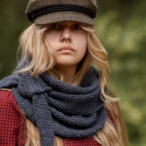 Könnte beinhalten: Eine Frau trägt einen grauen gehäkelten Schal und eine braune Tweed-Newsboy-Mütze mit schwarzen Seildetails. Die Frau trägt auch ein rot-schwarz kariertes Hemd. Der Hintergrund ist ein verschwommenes Grün.