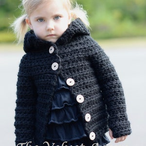May include: A black hooded cardigan with wooden buttons for a young child. The cardigan is crocheted and has a textured look. The child is wearing a black ruffled shirt and blue jeans.