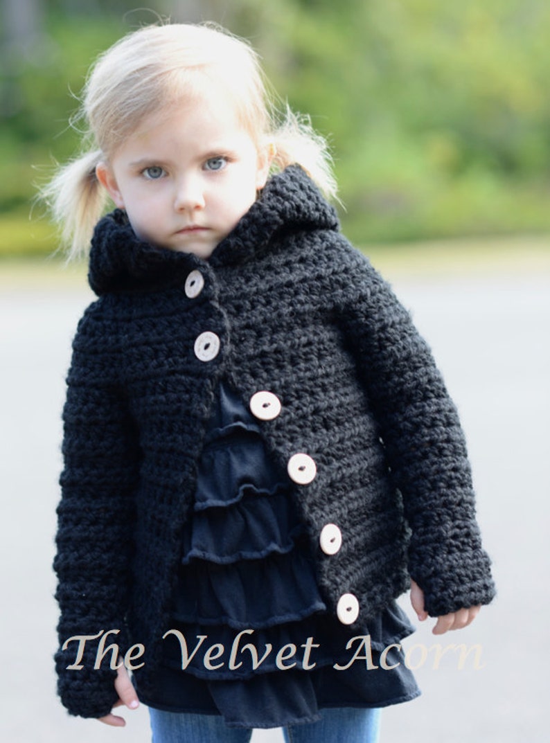 May include: A black hooded cardigan with wooden buttons for a young child. The cardigan is crocheted and has a textured look. The child is wearing a black ruffled shirt and blue jeans.