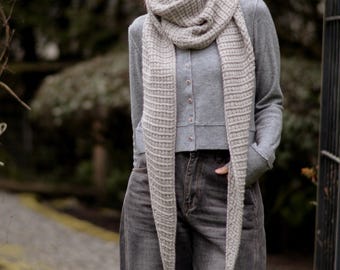 May include: A light gray knitted scarf with a textured pattern is draped around the neck and hangs down the front. The scarf is paired with a gray cardigan and dark gray jeans. The background is a blurred outdoor scene.