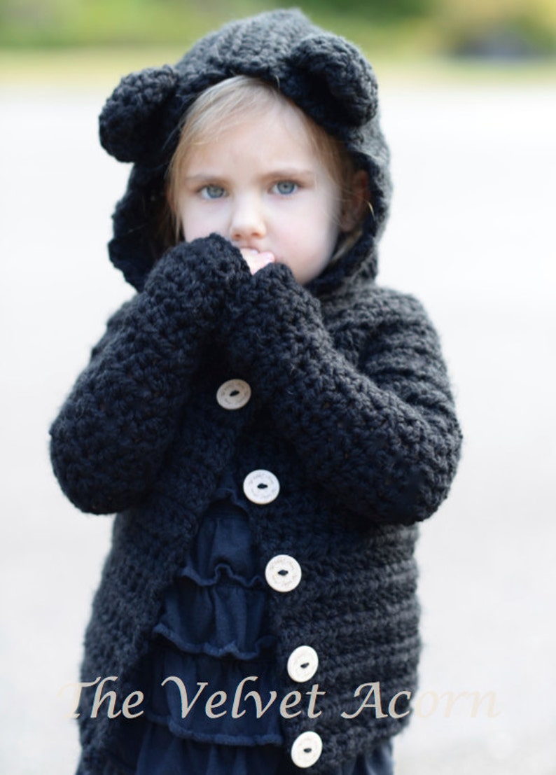 May include: A black crocheted hooded cardigan with bear ears, featuring wooden buttons and a ruffled navy blue dress underneath. The text "The Velvet Acorn" is visible at the bottom of the image.