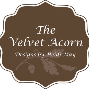 May include: A brown label with a white border and rounded edges. The text "The Velvet Acorn Designs by Heidi May" is written in white cursive font. A stylised acorn and leaf design is below the text.