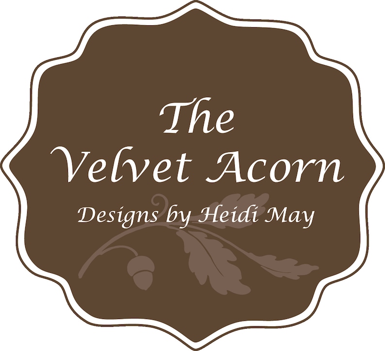 May include: A brown label with a white border and rounded edges. The text "The Velvet Acorn Designs by Heidi May" is written in white cursive font. A stylised acorn and leaf design is below the text.