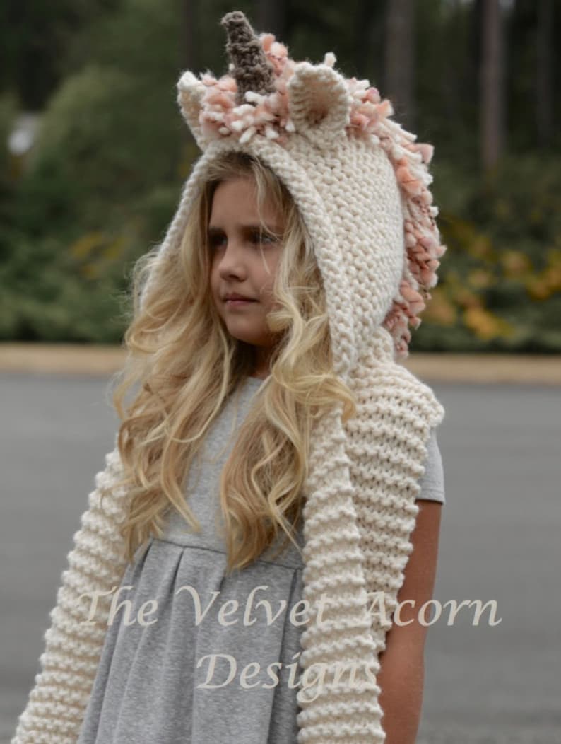 May include: A white knitted unicorn hat with a pink and brown horn and a long scarf. The hat has a fluffy pink trim around the hood and the scarf is knitted in a chunky stitch.