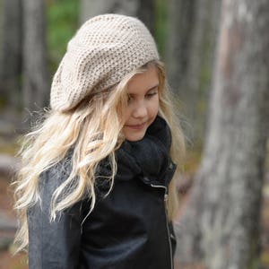 May include: A young girl with long blonde hair wearing a beige knitted beret and a black leather jacket. She is standing in a forest setting.