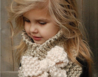 May include: A young girl wearing a brown and white knitted cowl with a large white knitted bow. The cowl is a warm and cozy accessory for the winter.