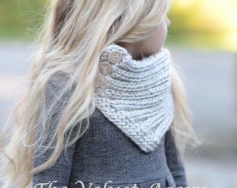 May include: A young girl wearing a gray dress with a white knitted scarf with wooden buttons. The scarf is tied around her neck and the buttons are visible on the front. The text "The Velvet Acorn Designs" is visible at the bottom of the image.