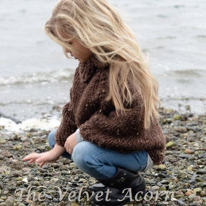 May include: A young girl with long blonde hair is wearing a brown knitted sweater and blue jeans. She is squatting on a rocky beach, looking down at the ground.