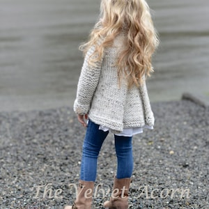 Pu&ograve; includere: Una bambina indossa un cardigan di maglia grigio chiaro con un motivo maculato, jeans blu e stivali marroni. Il cardigan ha una chiusura con bottone e un orlo leggermente svasato. La bambina &egrave; in piedi su una superficie di ghiaia. Il testo "The Velvet Acorn" &egrave; visibile nella parte inferiore dell'immagine.