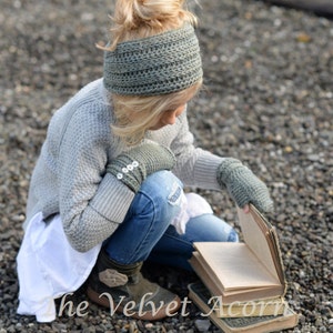 May include: A young person wearing a gray crocheted headband, a gray sweater, blue jeans, and gray crocheted fingerless gloves is sitting on a bed of gravel and reading a book. The book is open and the text on the cover reads "The Velvet Acorn".