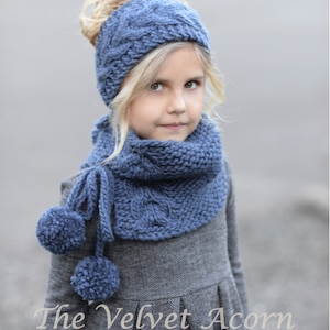 Peut inclure: Une jeune fille portant un bandeau et une écharpe en tricot bleu avec des pompons. L'écharpe est nouée autour de son cou et le bandeau est porté sur sa tête. Elle porte une robe grise à motif chevron. Le texte "The Velvet Acorn" est visible en bas de l'image.