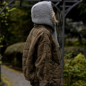 May include: A person wearing a gray knitted hooded scarf and a brown quilted jacket.
