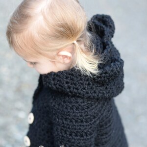 May include: A black crocheted hooded jacket with white buttons. The jacket is worn by a young child with blonde hair.