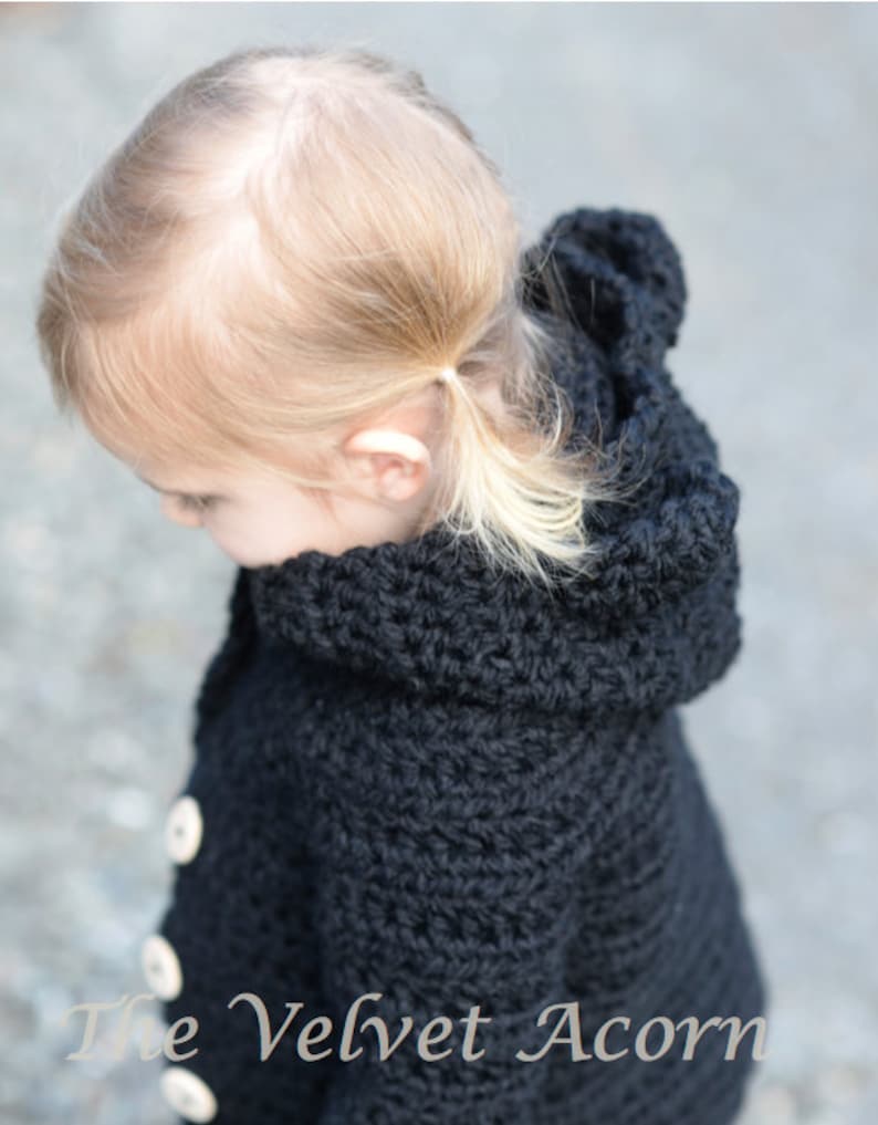 May include: A black crocheted hooded jacket with white buttons. The jacket is worn by a young child with blonde hair.