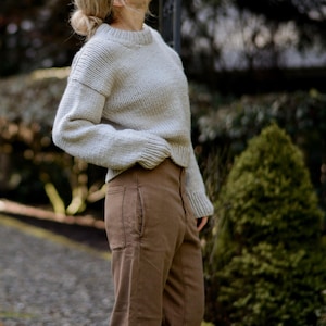 Puede incluir: Una mujer con un suéter de punto gris claro y pantalones marrones. El suéter tiene un corte holgado y un cuello redondo. Los pantalones son de pierna recta.