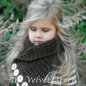 May include: A young girl wearing a brown knitted cowl neck scarf with white buttons. The scarf is made with a textured stitch and has a simple, elegant design. The girl has long blonde hair and is wearing a striped shirt. The text "The Velvet Acorn" is visible on the scarf.