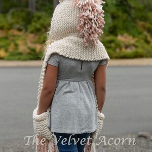 May include: A white knitted unicorn hooded scarf with a pink and white mane and tail. The scarf has pockets on the sides and long fringe.
