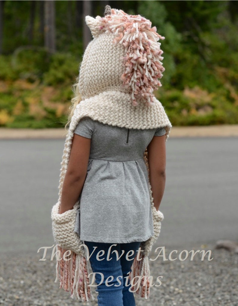 May include: A white knitted unicorn hooded scarf with a pink and white mane and tail. The scarf has pockets on the sides and long fringe.