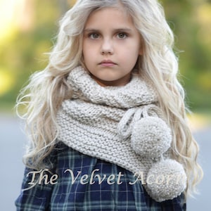 May include: A young girl with long blonde hair wearing a beige knitted scarf with two pom-poms. The scarf is draped around her neck and the pom-poms are hanging down. The girl is wearing a blue and black plaid shirt. The text "The Velvet Acorn" is visible in the bottom left corner of the image.