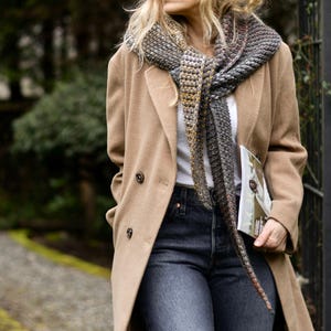 May include: A woman wearing a tan coat, dark wash jeans, and a patterned scarf. The scarf features shades of gray, brown, and orange. She is holding a magazine. The background is a blurred outdoor scene with trees and a pathway.