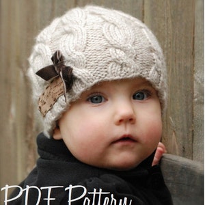 May include: A baby wearing a beige knitted hat with a brown bow and a leather patch. The hat has a cable knit pattern. The baby is looking at the camera.