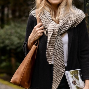 May include: A woman wearing a black coat and a patterned scarf in shades of brown and cream. She is carrying a brown leather tote bag and holding a magazine. The scarf is wrapped around her neck, and the bag is hanging from her shoulder.
