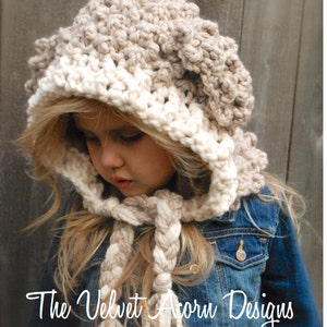 May include: A young girl wearing a white and brown crocheted hooded hat with a matching braided scarf. The hat has a textured, puffy design. The scarf has tassels at the ends. The girl is wearing a denim jacket.