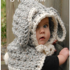 May include: A gray and white crocheted hooded scarf with bunny ears. The scarf has a white trim and a white pom-pom on the end. The scarf is being worn by a baby.