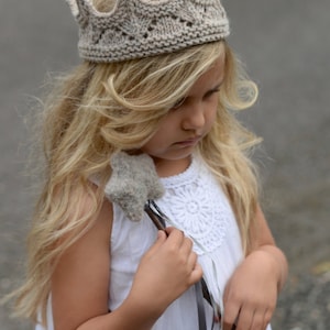 May include: A young girl wearing a knitted crown headband and a white dress holds a star-shaped wand. The wand is made of gray yarn and has a black ribbon tied around it. The girl's hair is long and blonde. The background is a blurry gray surface.