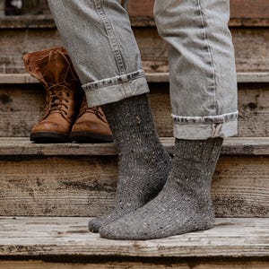 Peut inclure: Une paire de chaussettes en laine grise à motif moucheté. Les chaussettes sont portées par une personne portant un jean bleu et des bottes en cuir marron.